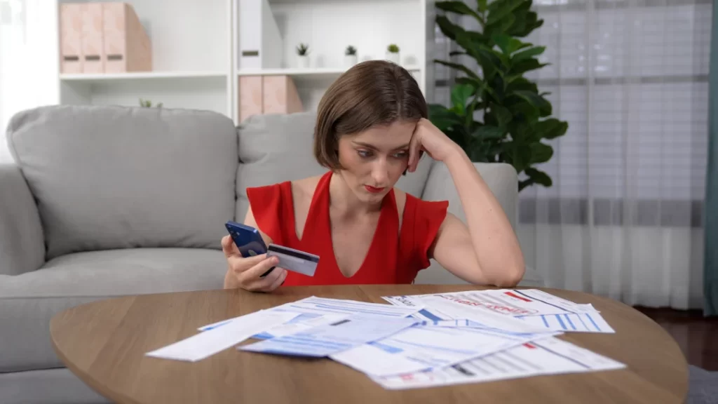 A woman seated at a table, examining bills while holding a credit card, reflecting on her financial situation.