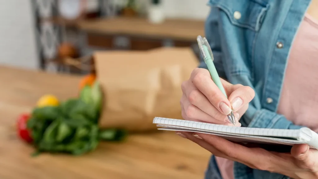 Woman writing in a notebook with a pen, surrounded by fresh vegetables in the background.