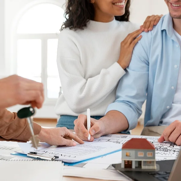 A couple sits at a table with a laptop and a house model, discussing plans for their future home.
