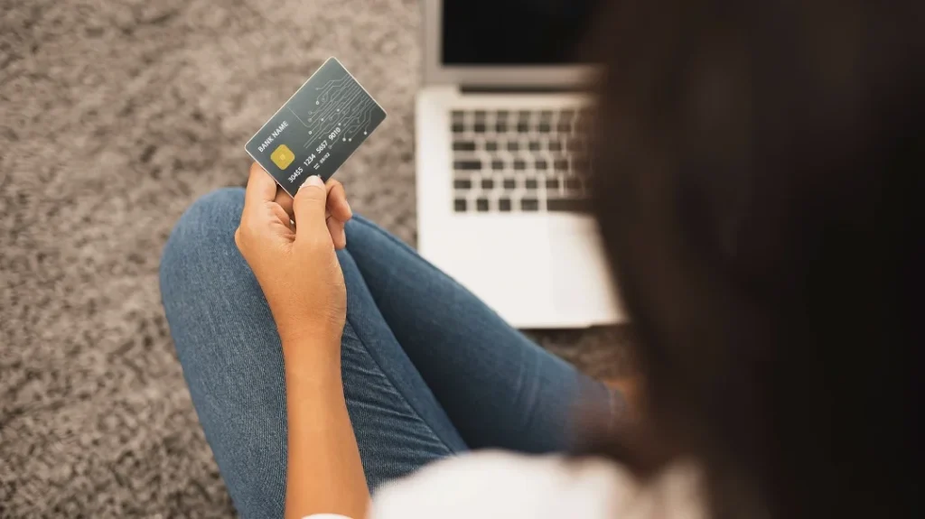 A woman sitting on the floor holds a credit card, looking thoughtful and engaged in her task.