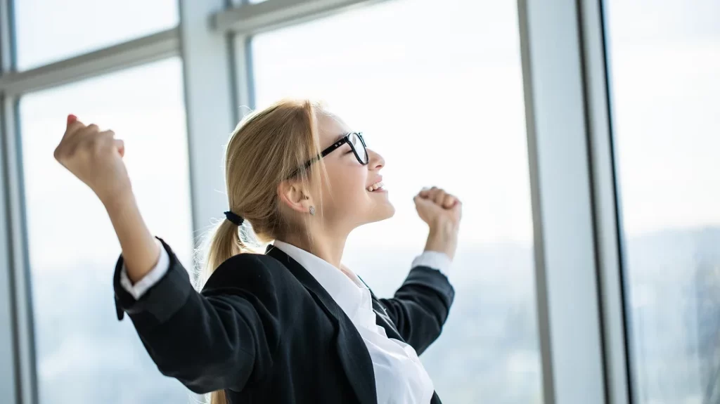 A woman in a business suit is raising her arms in a gesture of celebration or triumph.