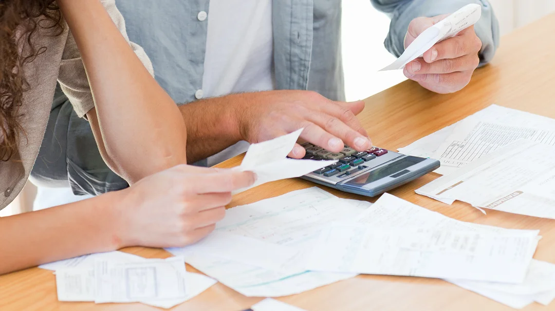 A man and woman are reviewing a calculator and financial papers together at a table.