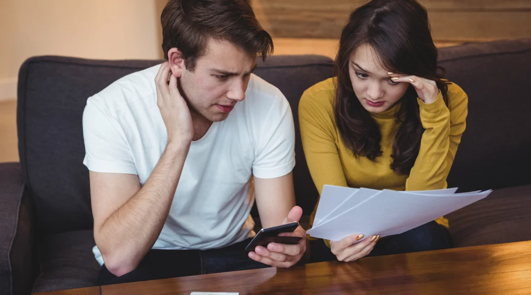 Couple sitting on a couch, reviewing financial documents, appearing concerned about their financial crisis. Couple sitting on a couch, reviewing financial documents, appearing concerned about their financial crisis.