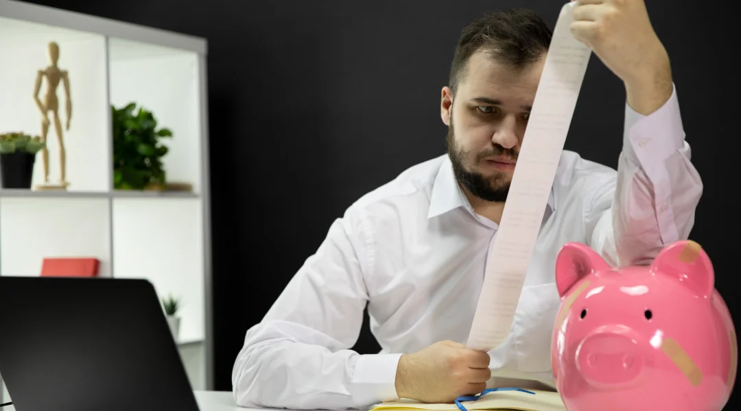 A man holds a piggy bank and a roll of paper, symbolizing his efforts to manage personal debts.